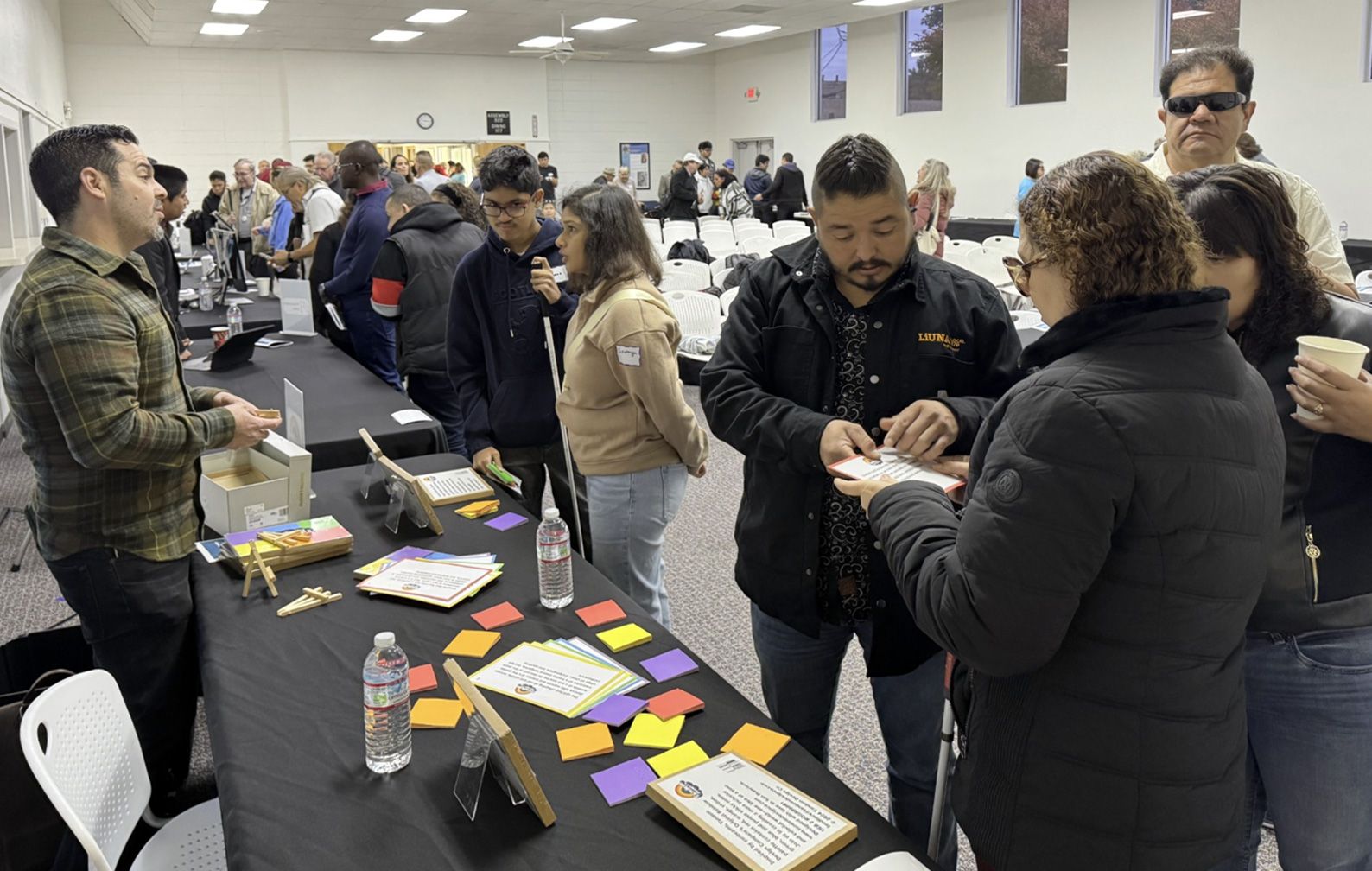 Tandem Design Co representative standing at their table with 6 guests visiting the table, engaged, and asking questions. Colorful braille sticky notes lay spread out on the table. Behind the table are other PitchAbility entrepreneur tables lined up around the border of the auditorium.