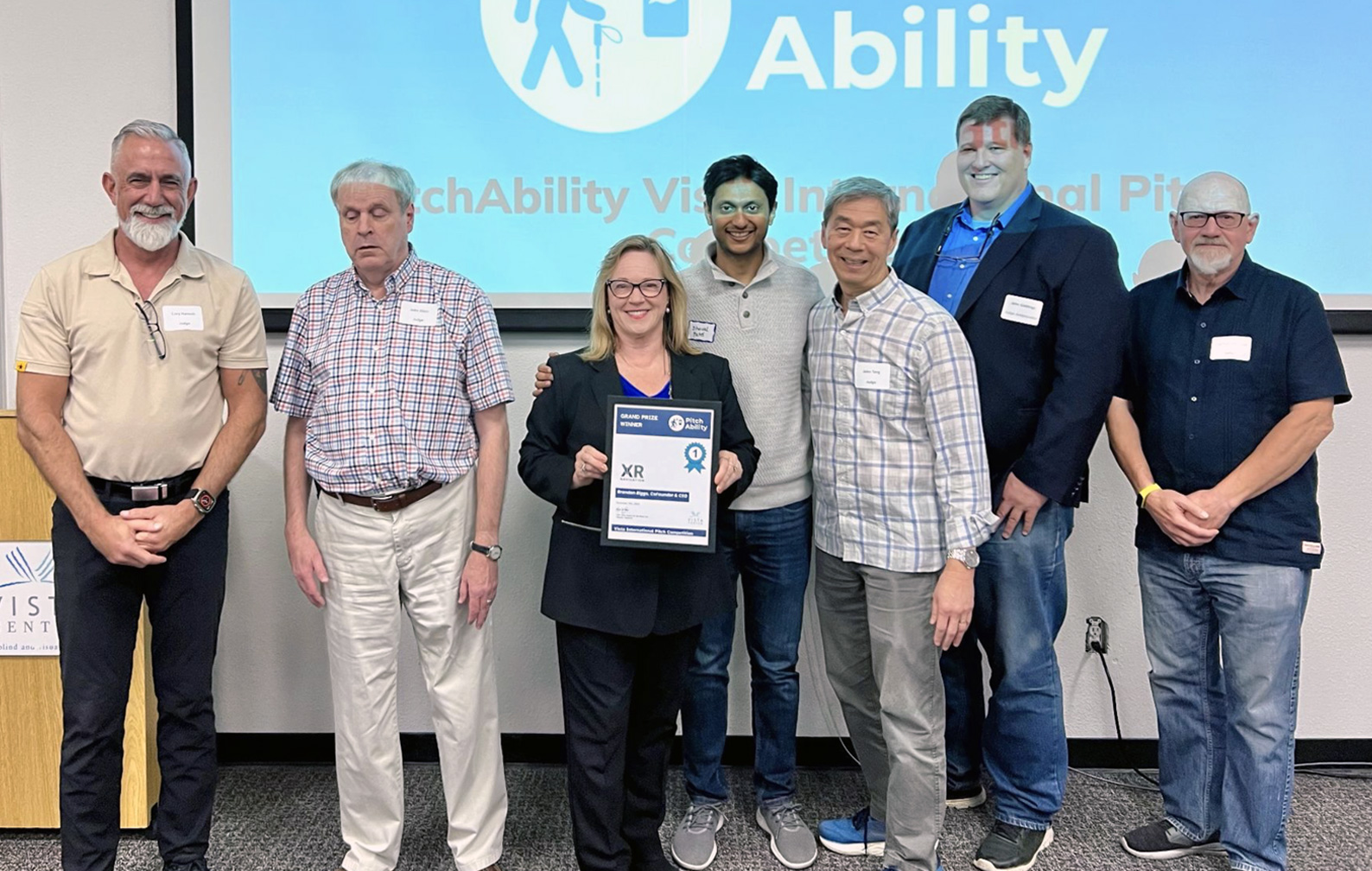 PitchAbility 2025 Finalist and Judge photo, including Cory Hanosh (President at Northstate AT), John Glass (Former Vista Center Chair of the Board), Karae Lisle (Vista Center CEO), Dhaval Patel (CEO at Lotus), John Tang (Accessibility Research Leader at Microsoft), John Giddings (PitchAbility 20205 emcee), and Spencer Johnson (Former Technical Manager at Amazon). The XR Navigation framed winner's certificate is held in the front and center of the photo.